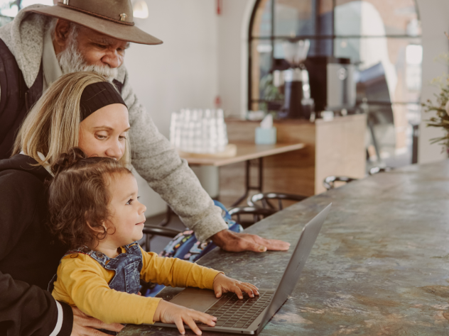 Photo of three generations of people sitting together, looking at a laptop on the counter of a cafe or restaurant.