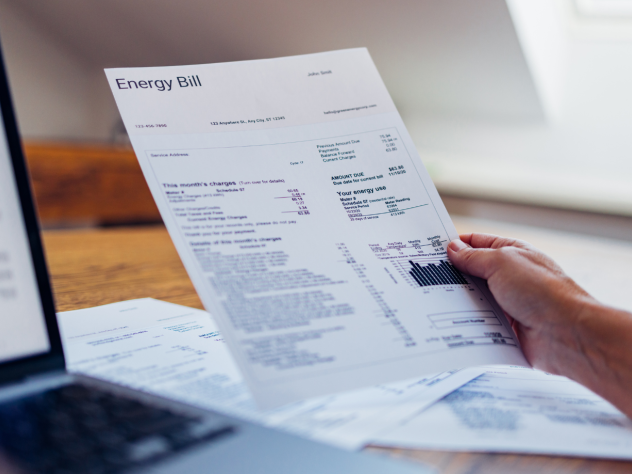 Photo of a hand holding a piece of paper with 'Energy Bill' written on it; next to a desktop and laptop.