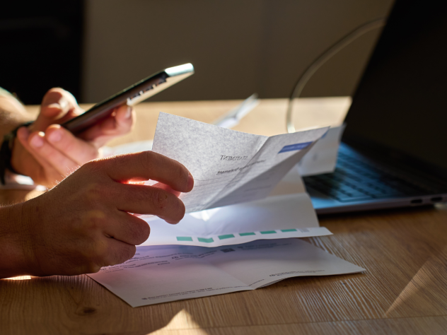 Photo of hands at a desk - one hand is holding a phone and the other is sorting through receipts/bills. There is also a laptop atop the desk.
