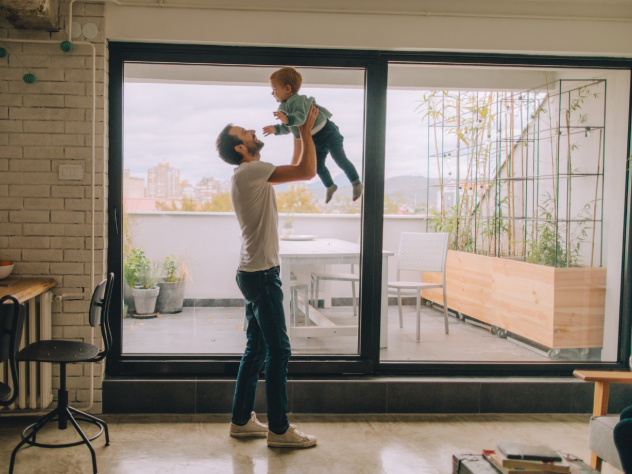 Photo of an interior apartment space, in which a parent is holding their child up in the air in a lounge room setting. Behind them, there is a sliding glass dooe leading to an external balcony area, with some potted plants and a seating area.