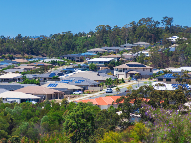 Photo of Australian rooftops amidst bushland, some with solar panels and set against a bright blue sky.