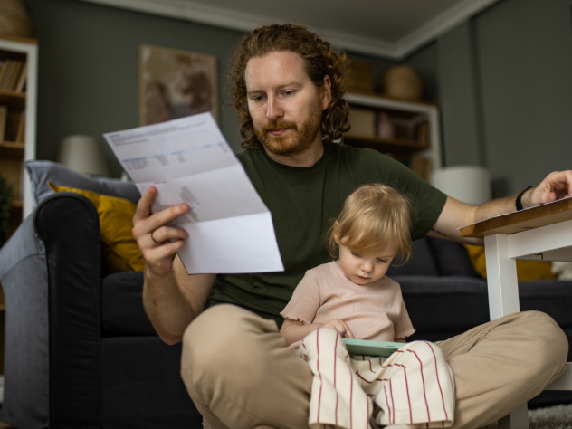 Photo of a parent with a child in their lap, sitting in a living area, looking at a white paper bill with a concerned expression.