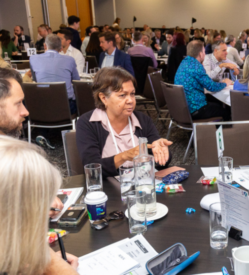 Photo of people in discussion indoors, sitting at a table, looking at the person who is talking.