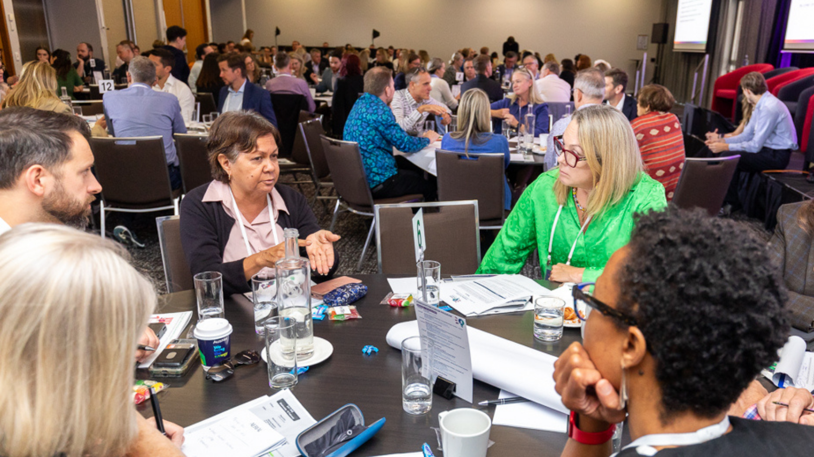 Photo of people in discussion indoors, sitting at a table, looking at the person who is talking.