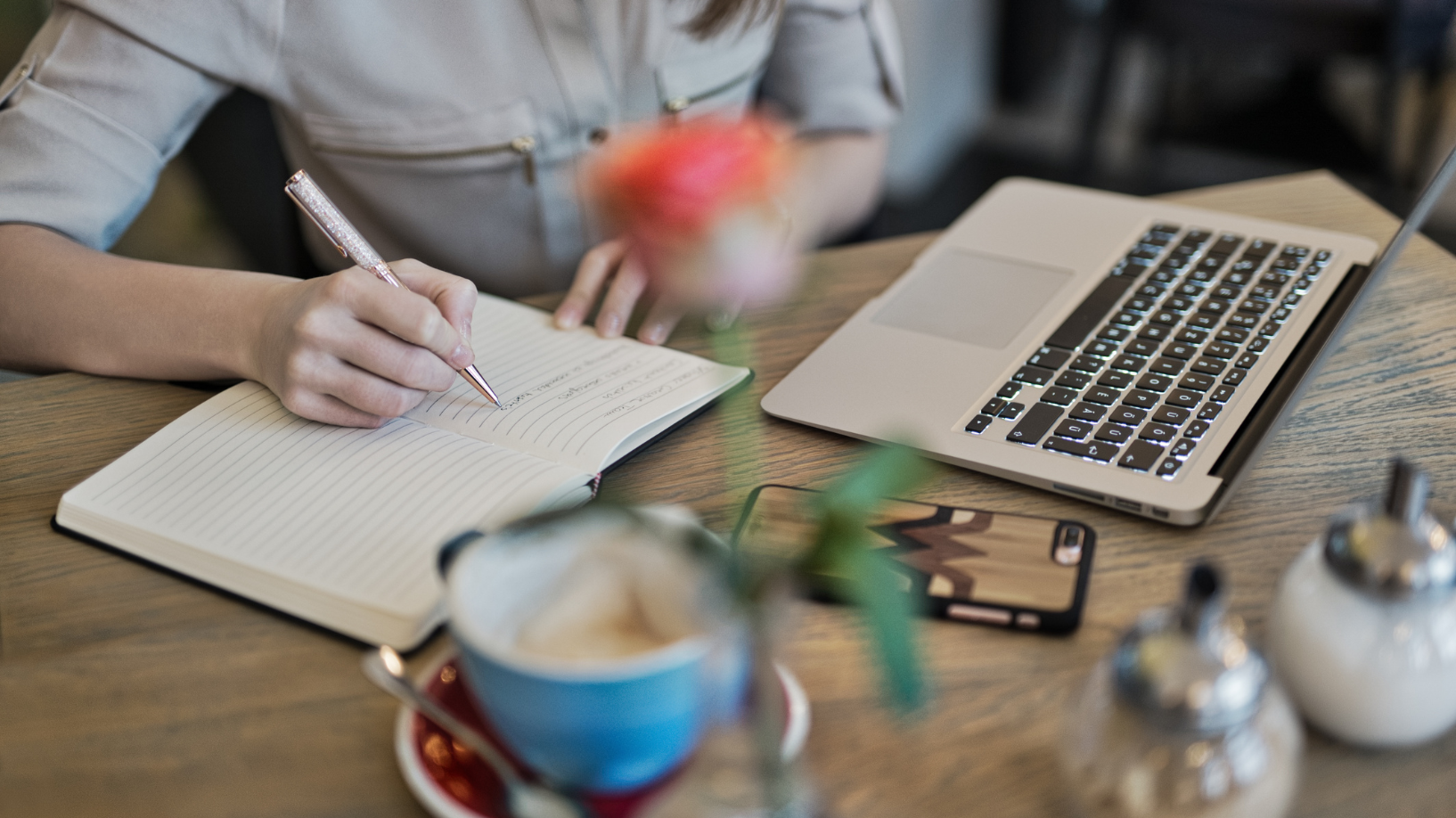 Photo of a person's hands on a desk, writing in a book. There is a cup of coffee, a sugar pot in the foreground, and an iPhone and a laptop in the middle ground.