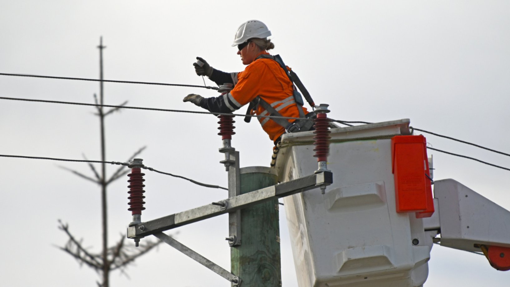 Photo of an electrical tradesperson fixing a transmission line from a cherry picker in hi vis, in front of an overcast sky