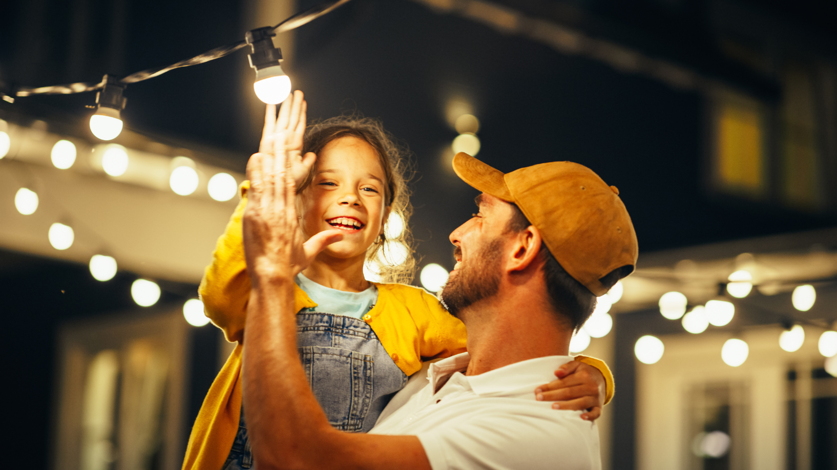 Photo of a dad and a daughter high-fiving underneath fairy lights, outdoors