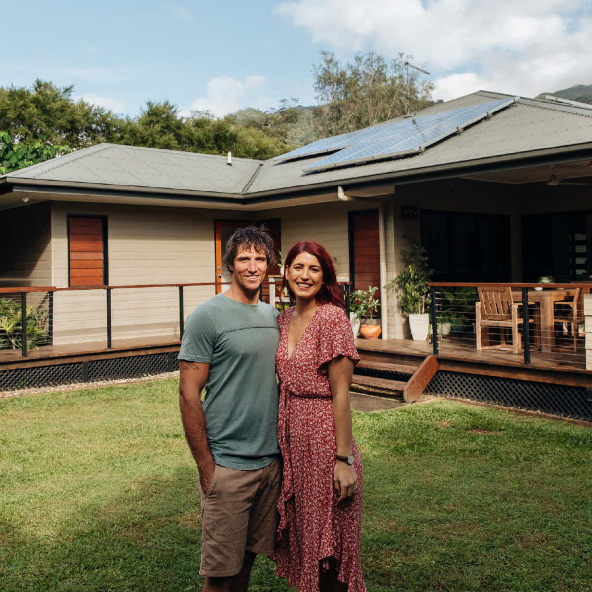 Photo of two people standing in front of their home smiling at the camera; their home has a big wraparound deck and solar panels on the rooftop, and is surrounded by trees and bright green grass