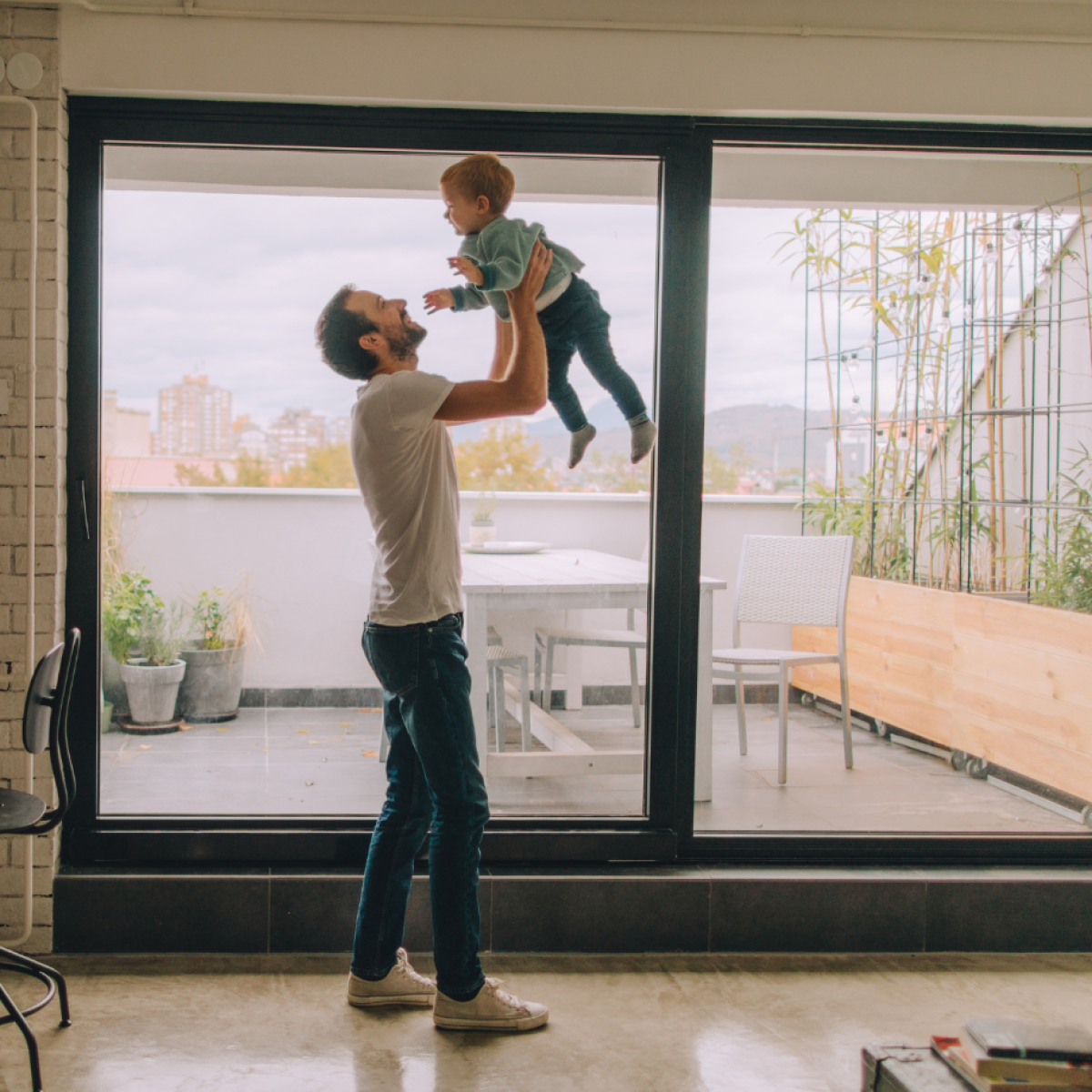 Photo of an interior apartment space, in which a parent is holding their child up in the air in a lounge room setting. Behind them, there is a sliding glass dooe leading to an external balcony area, with some potted plants and a seating area.