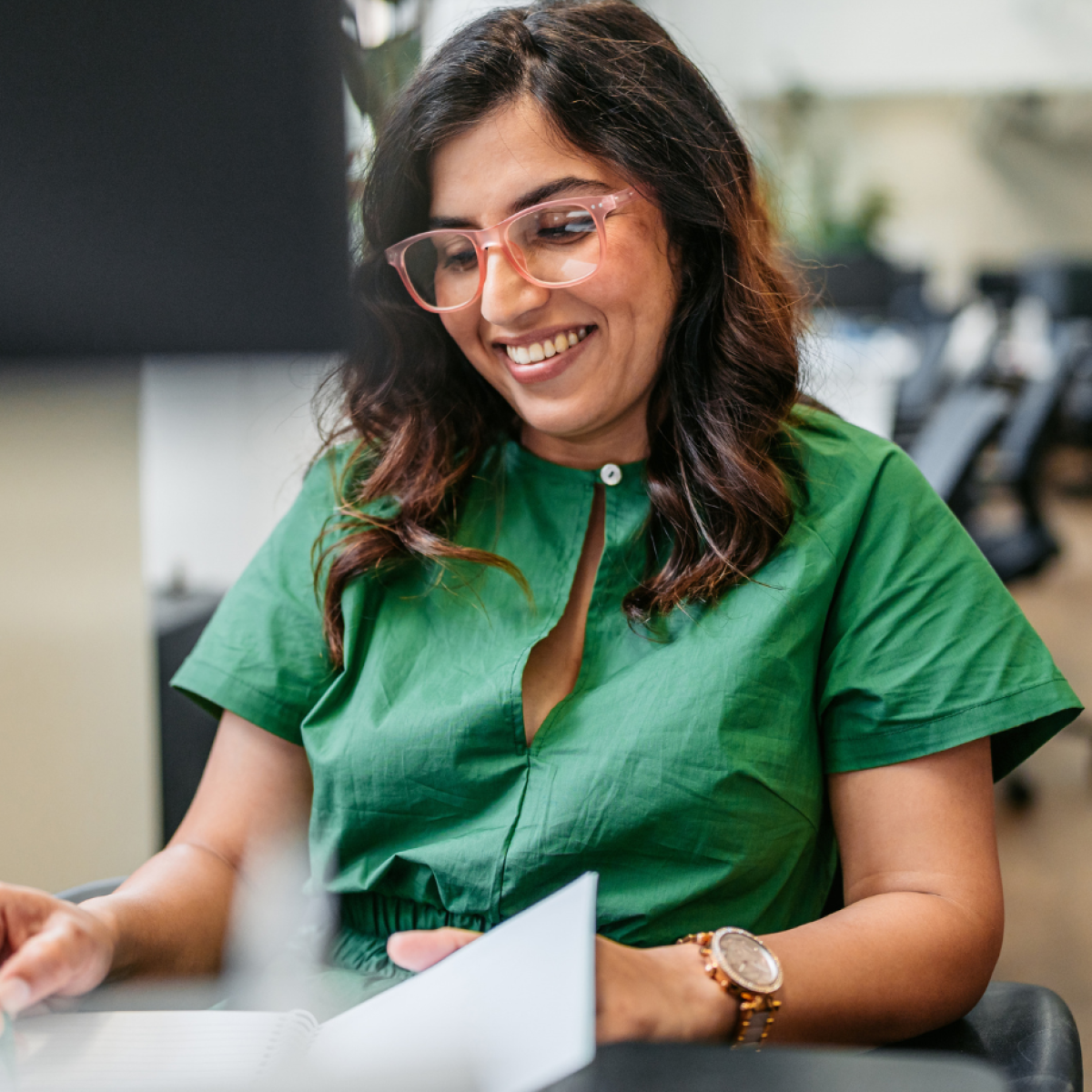 Photo of person smiling, wearing green shirt and holding green pen, seated at desk writing on notepad in office setting.