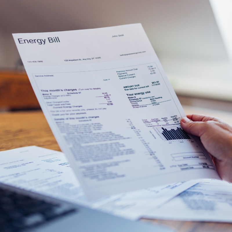 Photo of a hand holding a piece of paper with 'Energy Bill' written on it; next to a desktop and laptop.