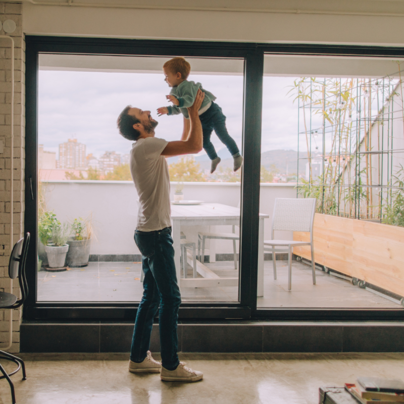 Photo of an interior apartment space, in which a parent is holding their child up in the air in a lounge room setting. Behind them, there is a sliding glass dooe leading to an external balcony area, with some potted plants and a seating area.