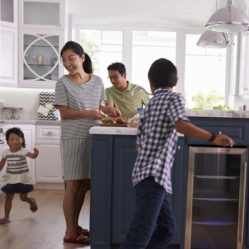 Photo of a family of four in a kitchen. The two kids are chasing each other around a kitchen island while the parents look on. Everyone looks quite happy.