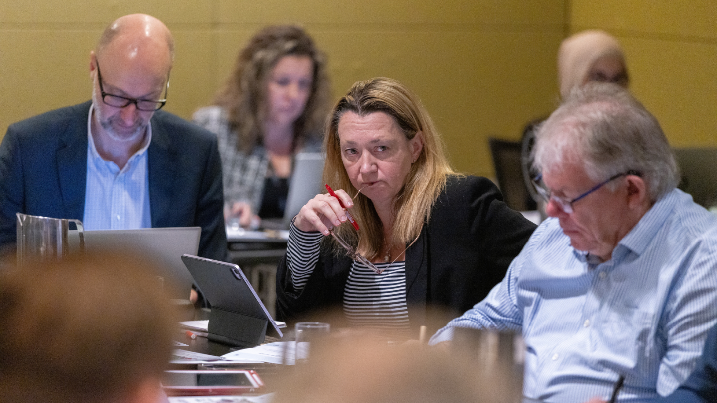 Photo of a person amidst a group of people, thinking, with one end of their glasses at their mouth