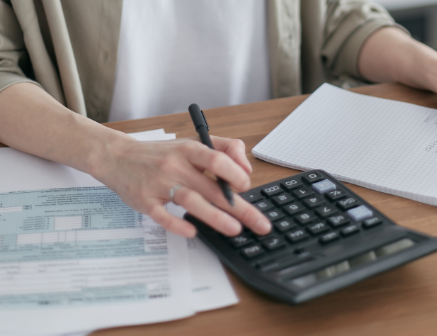Photo of a person's hands, holding a pen and typing into a calculator, next to some papers on a desk.