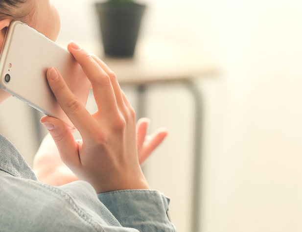 Photo of the back of a person's head, they are holding a mobile phone to their ear and talking into it. They are wearing a blue shirt.