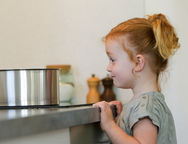 Photo of a girl with her hands on a kitchen counter, looking at a steel saucepan on an induction cooktop that is about eye height