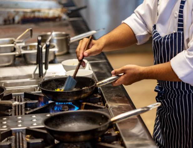 Close-up photo of a cook's arms stirring something in a frypan in a commercial kitchen, over a gas stove.