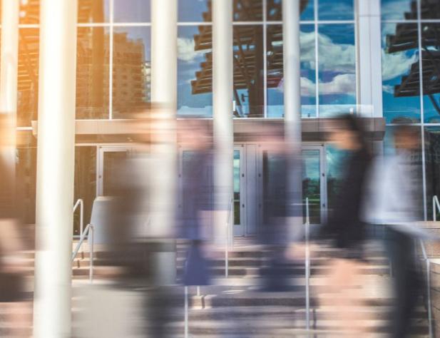Photo of an office building reflecting the sky and trees, with blurry figures walking in front of it. The blur looks intended to convey fast movement.