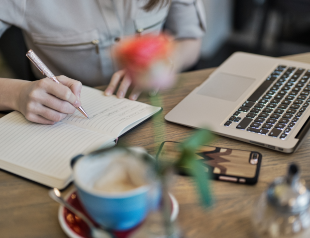 Photo of a person's hands on a desk, writing in a book. There is a cup of coffee, a sugar pot in the foreground, and an iPhone and a laptop in the middle ground.