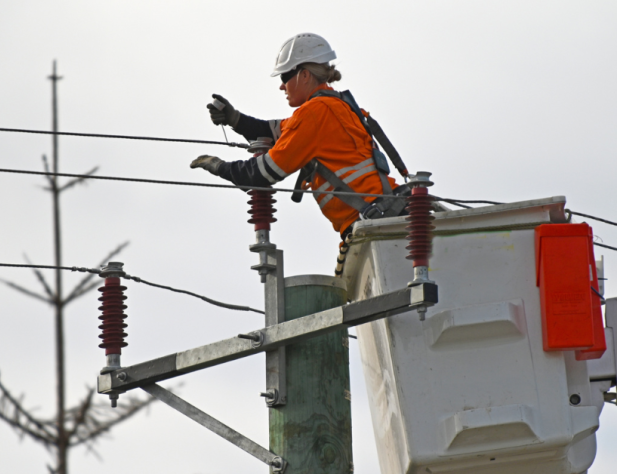 Photo of an electrical tradesperson fixing a transmission line from a cherry picker in hi vis, in front of an overcast sky