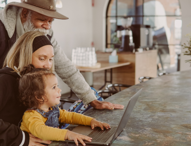 Photo of three generations of people sitting together, looking at a laptop on the counter of a cafe or restaurant.