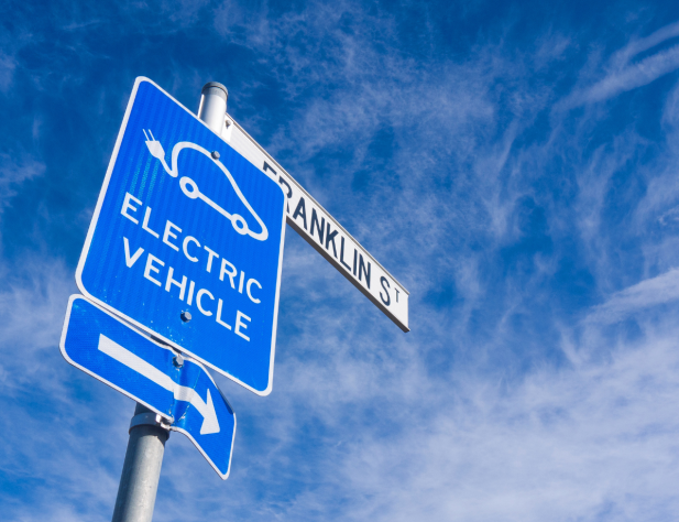 Photo of a blue sign against a blue sky with 'electric vehicle' written on it, in front of a street sign saying 'Franklin St'