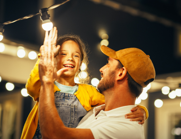 Photo of a dad and a daughter high-fiving underneath fairy lights, outdoors