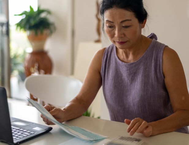 Photo of person in purple singlet top, looking at papers and using a calculator and their laptop, on a benchtop.