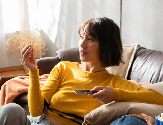 Photo of a person sitting in front of a couch in a yellow jumper, holding a piece of popcorn and a TV remote, looking at the TV.