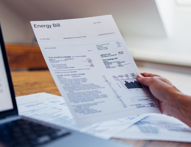Photo of a hand holding a piece of paper with 'Energy Bill' written on it; next to a desktop and laptop.