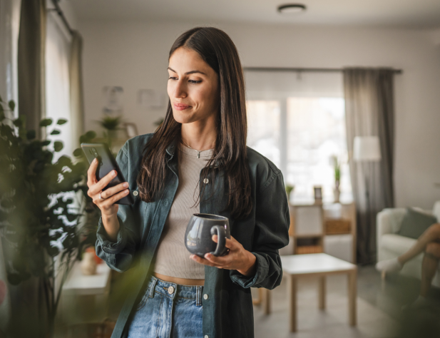 Photo of a person using a phone and holding a mug, standing next to a window. In the background is their living room.