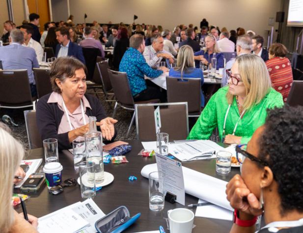 Photo of people in discussion indoors, sitting at a table, looking at the person who is talking.