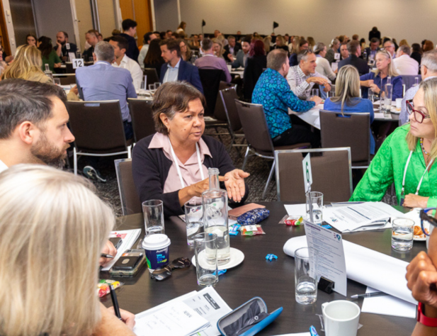 Photo of people in discussion indoors, sitting at a table, looking at the person who is talking.