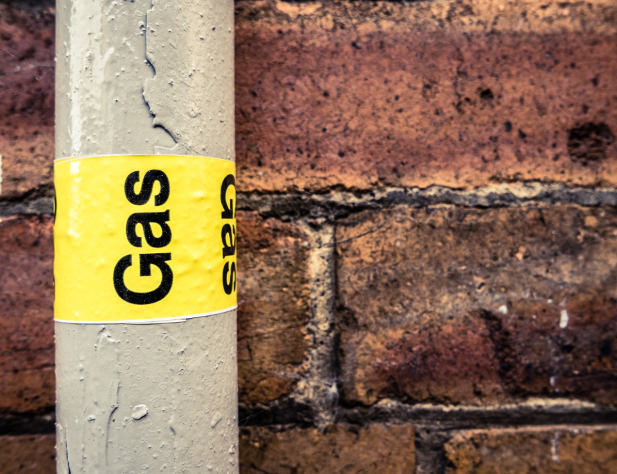 Close-up photo of a grey pipe with yellow tape and the word 'gas' on it, against a brick wall