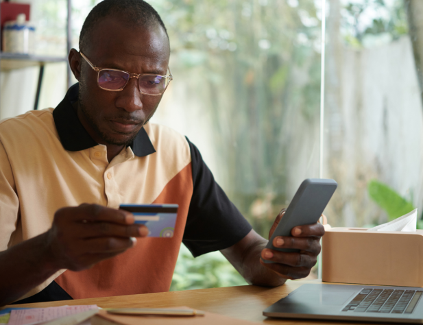Photo of a person looking at a credit card while also holding their phone, sitting at a desk with a laptop on it.