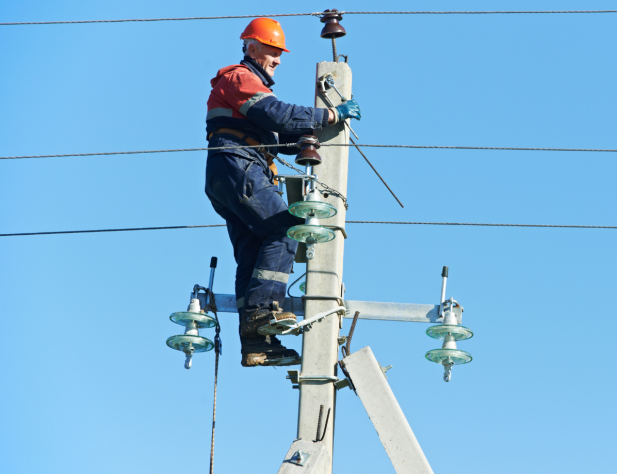 Photo of an electrical tradesperson in a hard hat, standing on a power pole against a bright blue sky