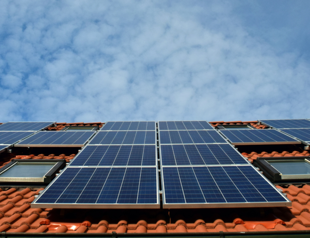 Photo of a terracotta tiled rooftop with solar panels on it, against a blue sky with wispy clouds.