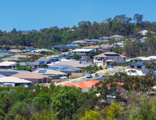 Photo of Australian rooftops amidst bushland, some with solar panels and set against a bright blue sky.