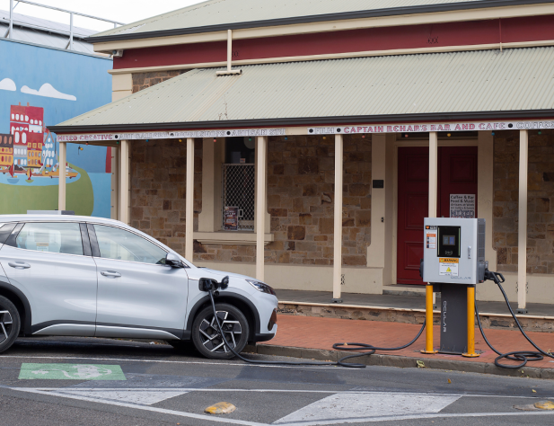 Photo of a car charging kerbside on a street in Port Adelaide, South Australia. The white car is connected to an electric vehicle charger situated on the footpath, in front of a shop.