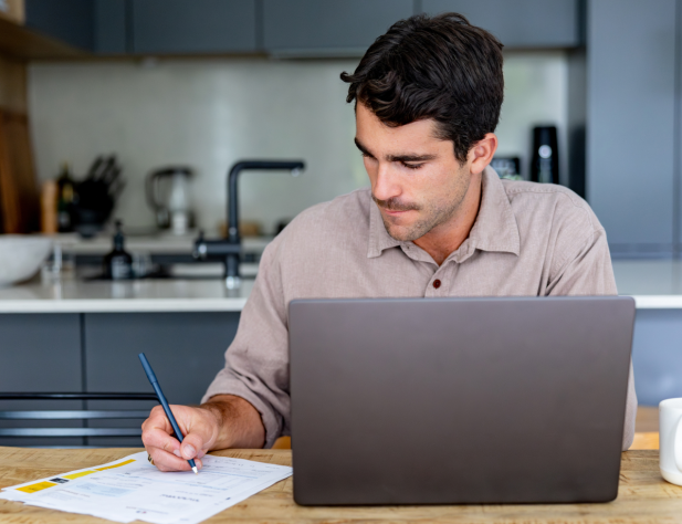 Photo of a person sitting at a desk writing something, with a laptop and coffee cup, against an interior kitchen.