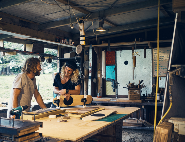 Photo of two people standing in a skateboard workshop space, surrounded by tools in their workspace and against a rural backdrop.