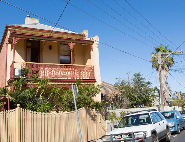 Photo of a cream-coloured terrace house in St Kilda, Victoria, on a street lined with parked cars and with power lines and poles, and a palm tree set against a bright blue sky.