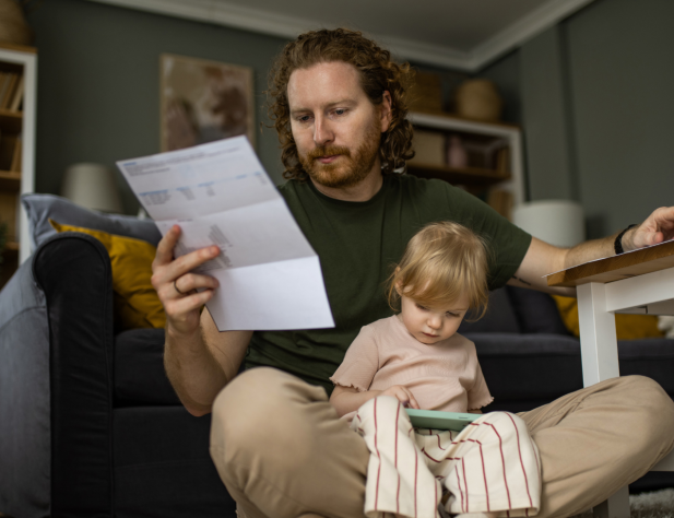 Photo of a parent with a child in their lap, sitting in a living area, looking at a white paper bill with a concerned expression.
