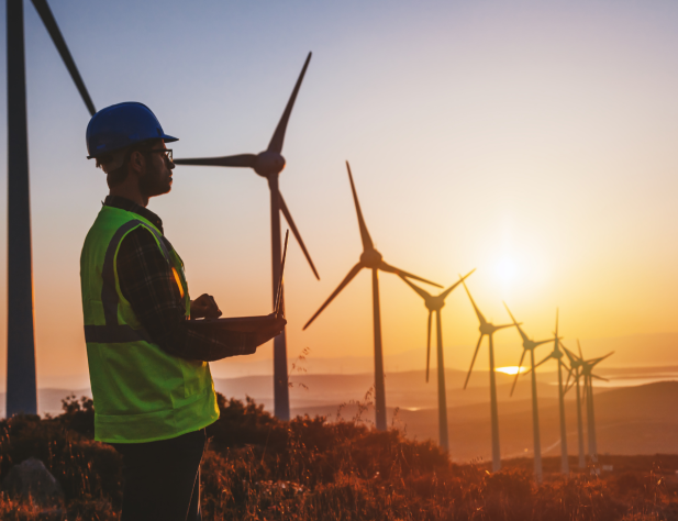 Photo of a person in a hard hat and hi vis vest standing next to wind turbines at sunset, looking into the distance.