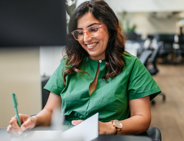 Photo of person smiling, wearing green shirt and holding green pen, seated at desk writing on notepad in office setting.