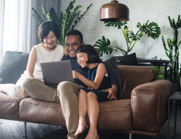 Photo of a family sitting around a laptop on a couch; they're smiling at what is on the laptop and there is a lamp and plants in the background