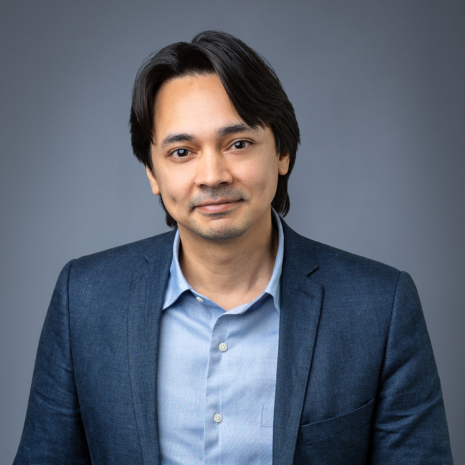 Photo of a person in a light blue shirt with a navy blue jacket, looking directly at the camera and smiling.