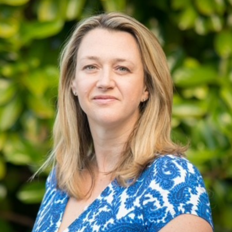 Photo of a person with medium length, light coloured hair, wearing a blue and white dress, in front of a leafy background.