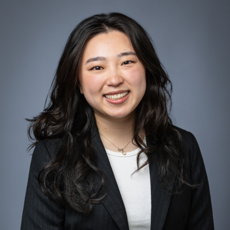 Photo of a person with long dark hair, smiling at the camera. They are wearing a white t-shirt and black suit jacket.